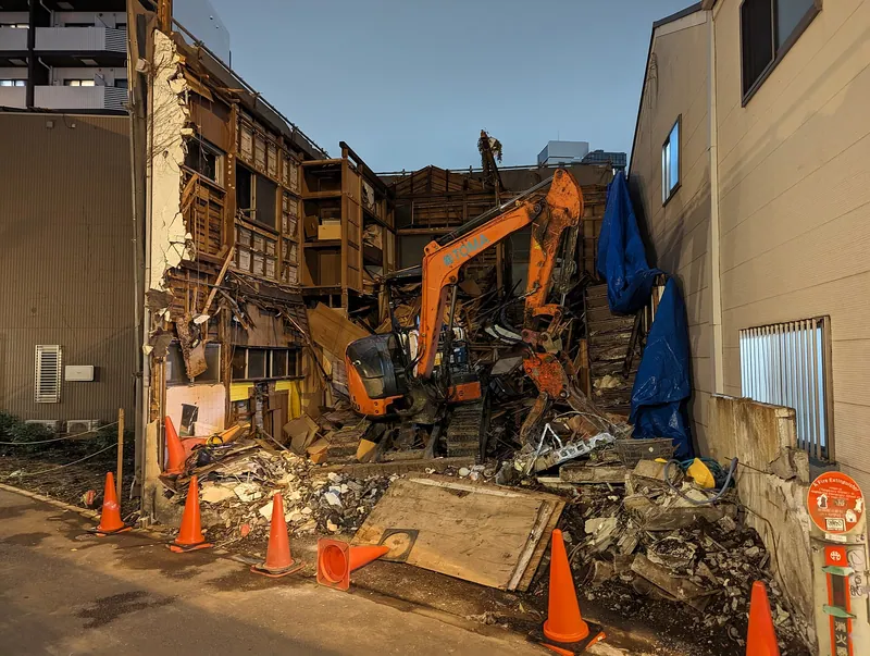 Casa antigua de madera a medio demoler, con una excavadora entre los escombros y conos de seguridad en la vereda